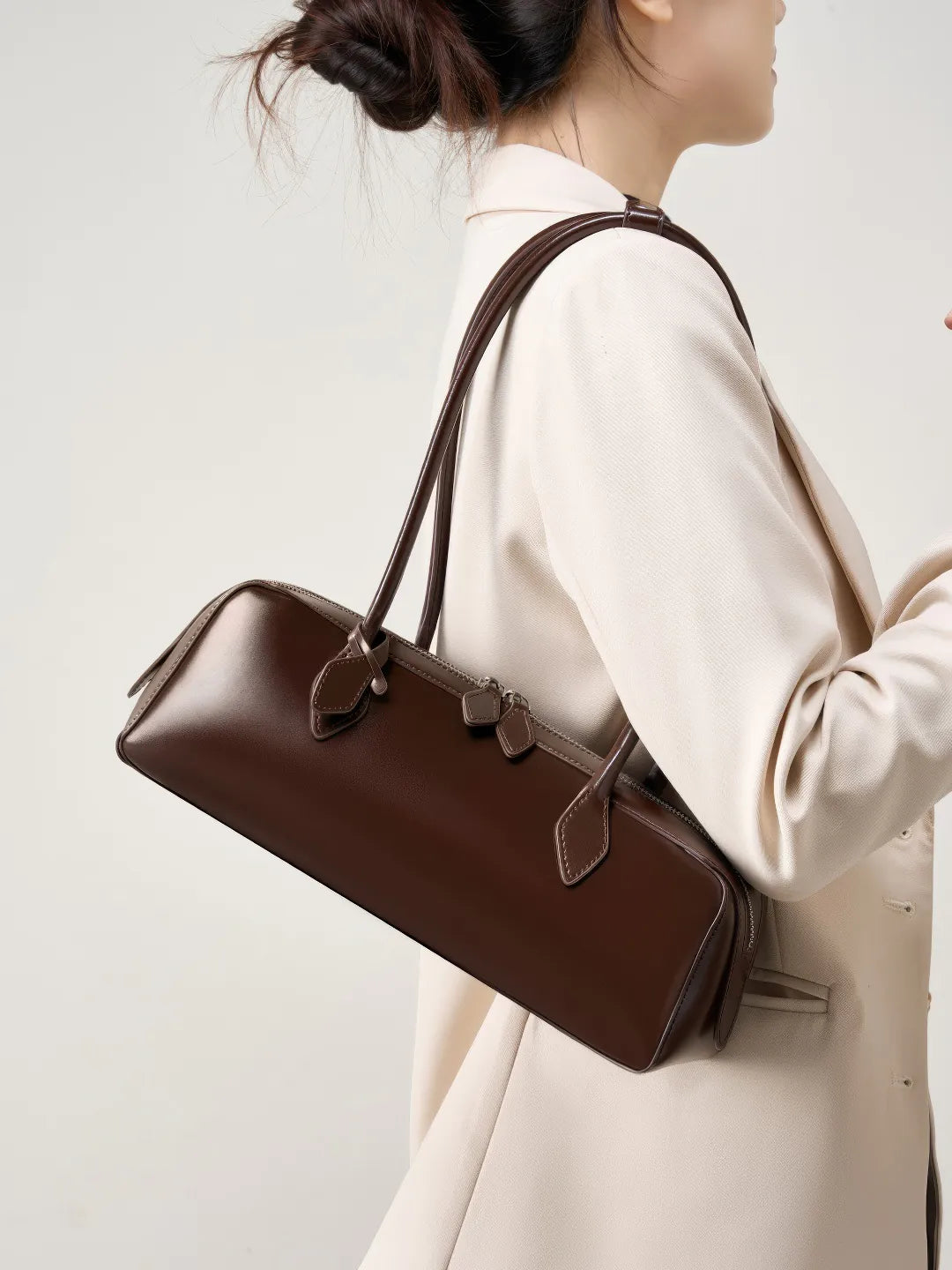 Brown leather handbag held by a person wearing a beige coat against a light background #Color_Brown