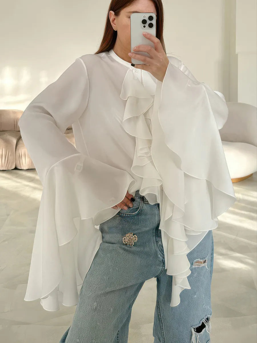 Person wearing a white ruffled blouse and light blue jeans indoors. #Color_White