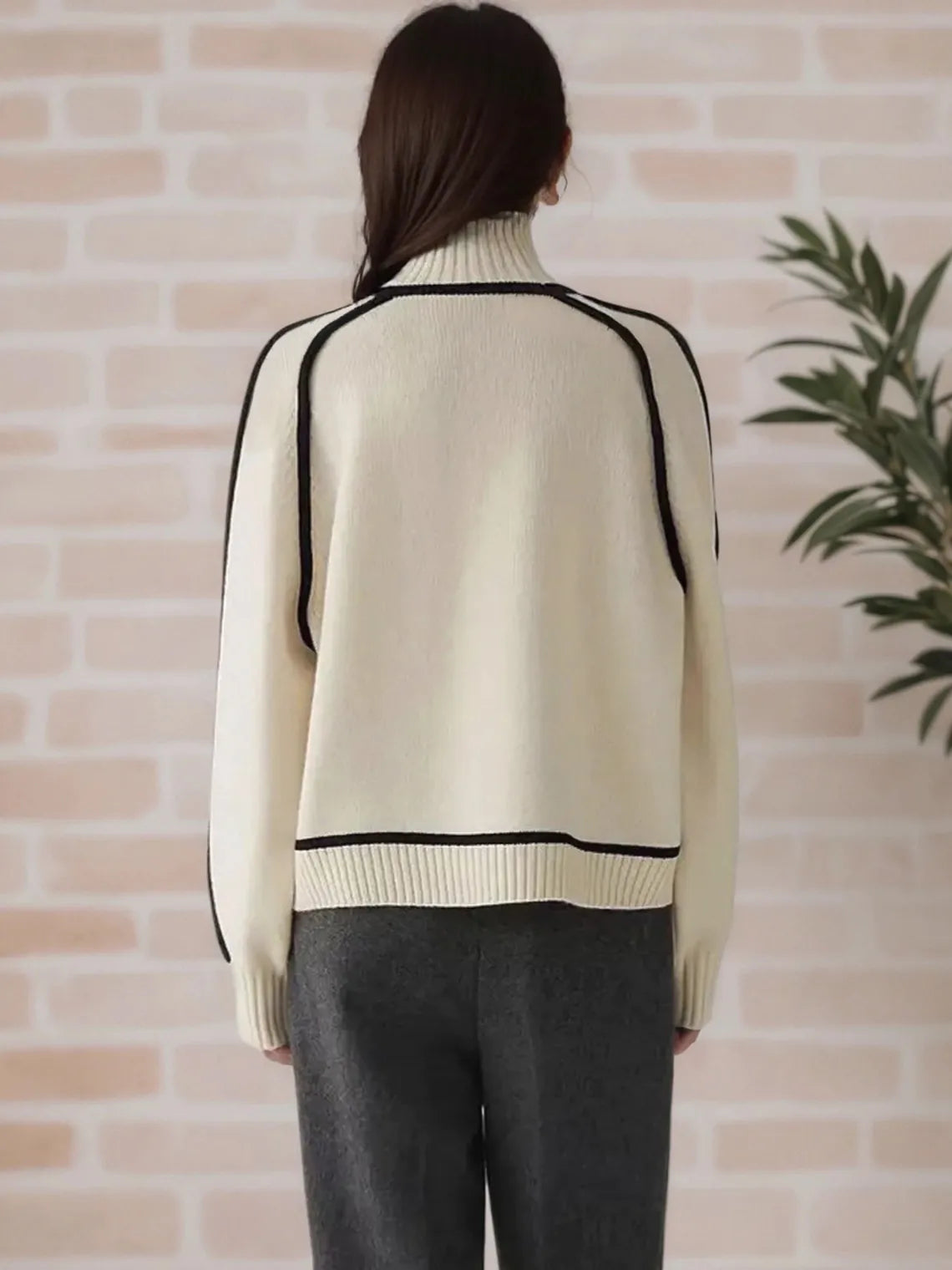 Person wearing a beige sweater with black trim against a brick wall. #Color_Beige