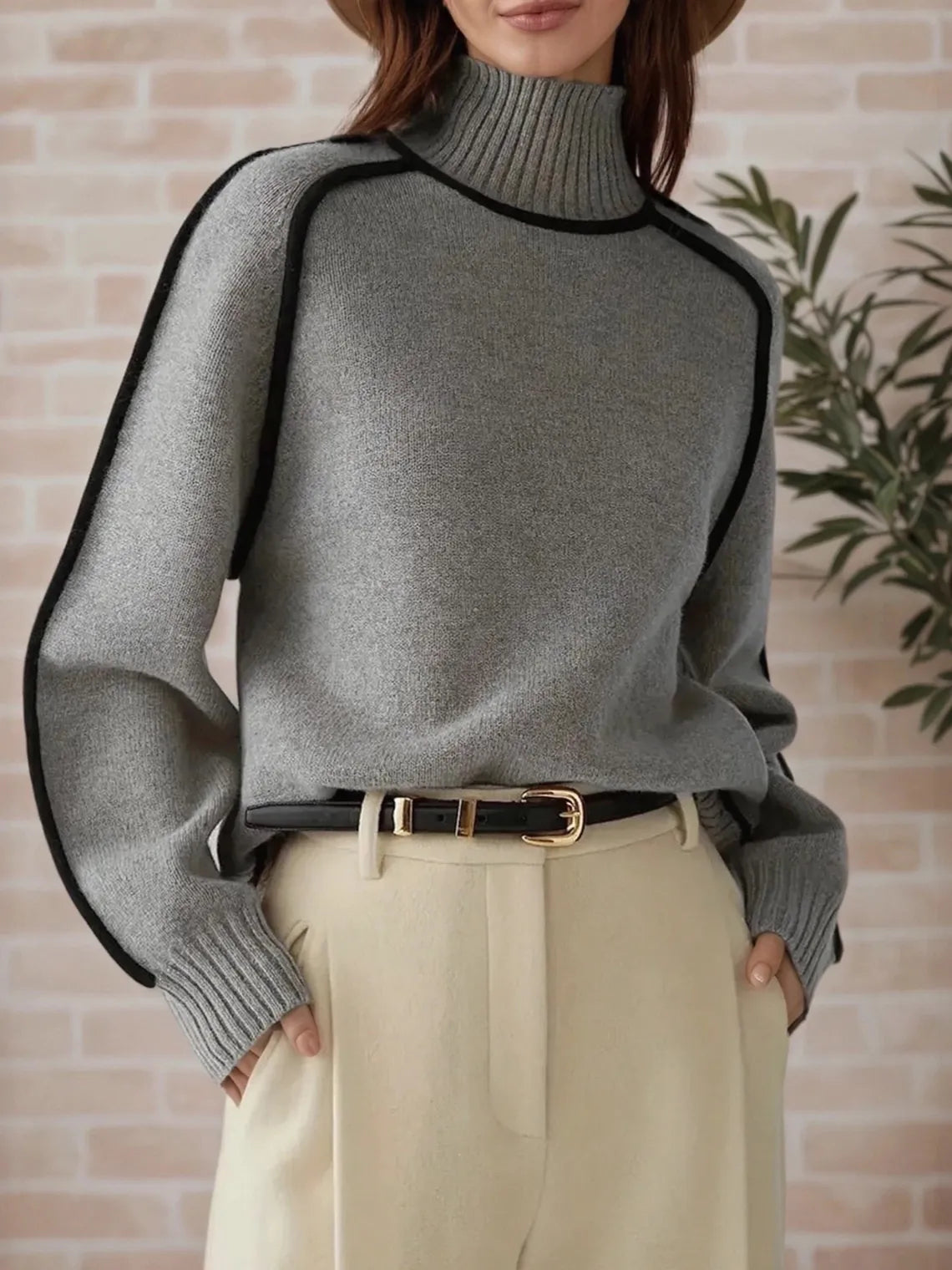 Person wearing a gray sweater with black trim against a brick wall. #Color_Gray