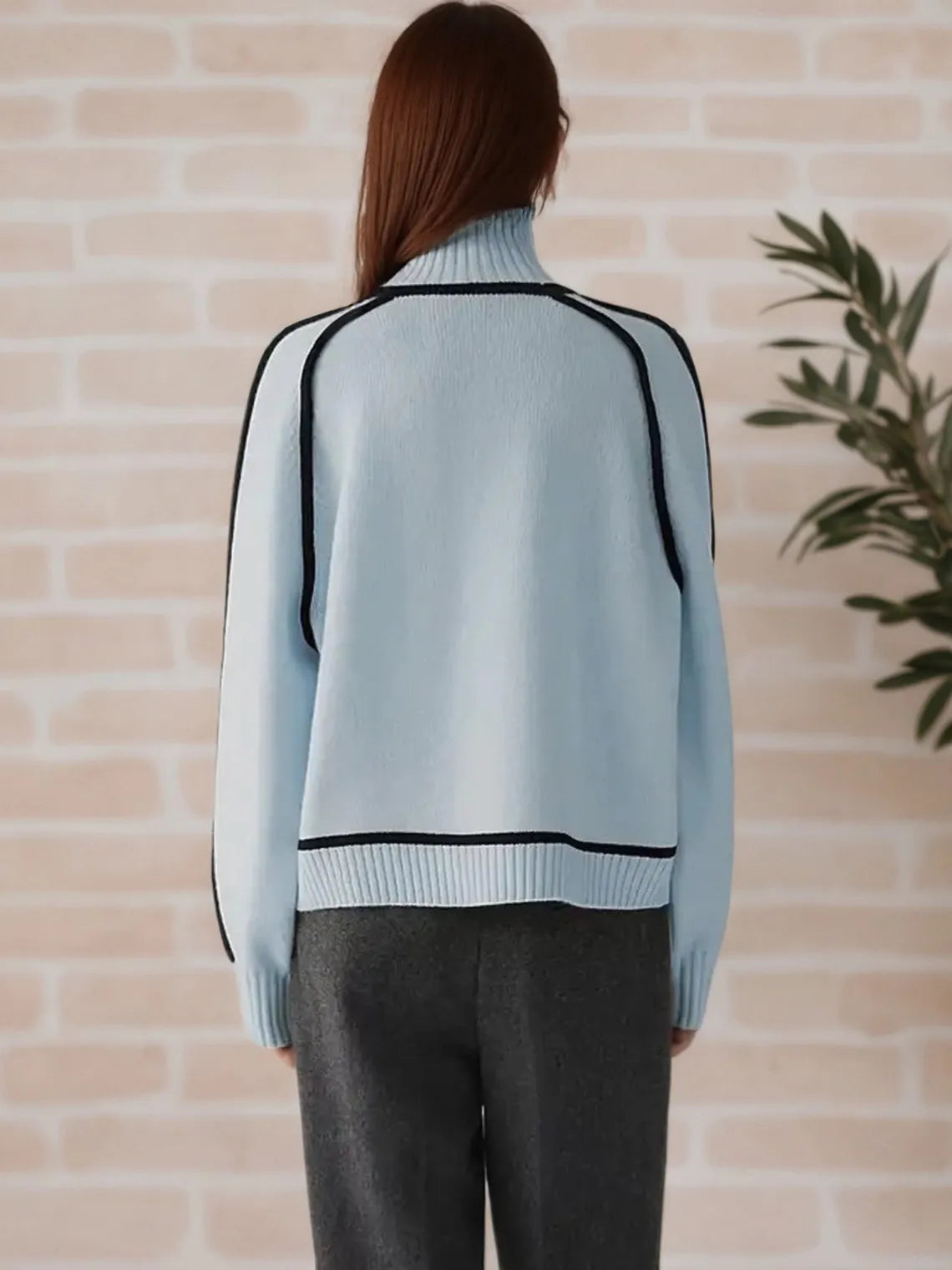 Person wearing a light blue sweater with black trim against a beige brick wall. #Color_LightBlue