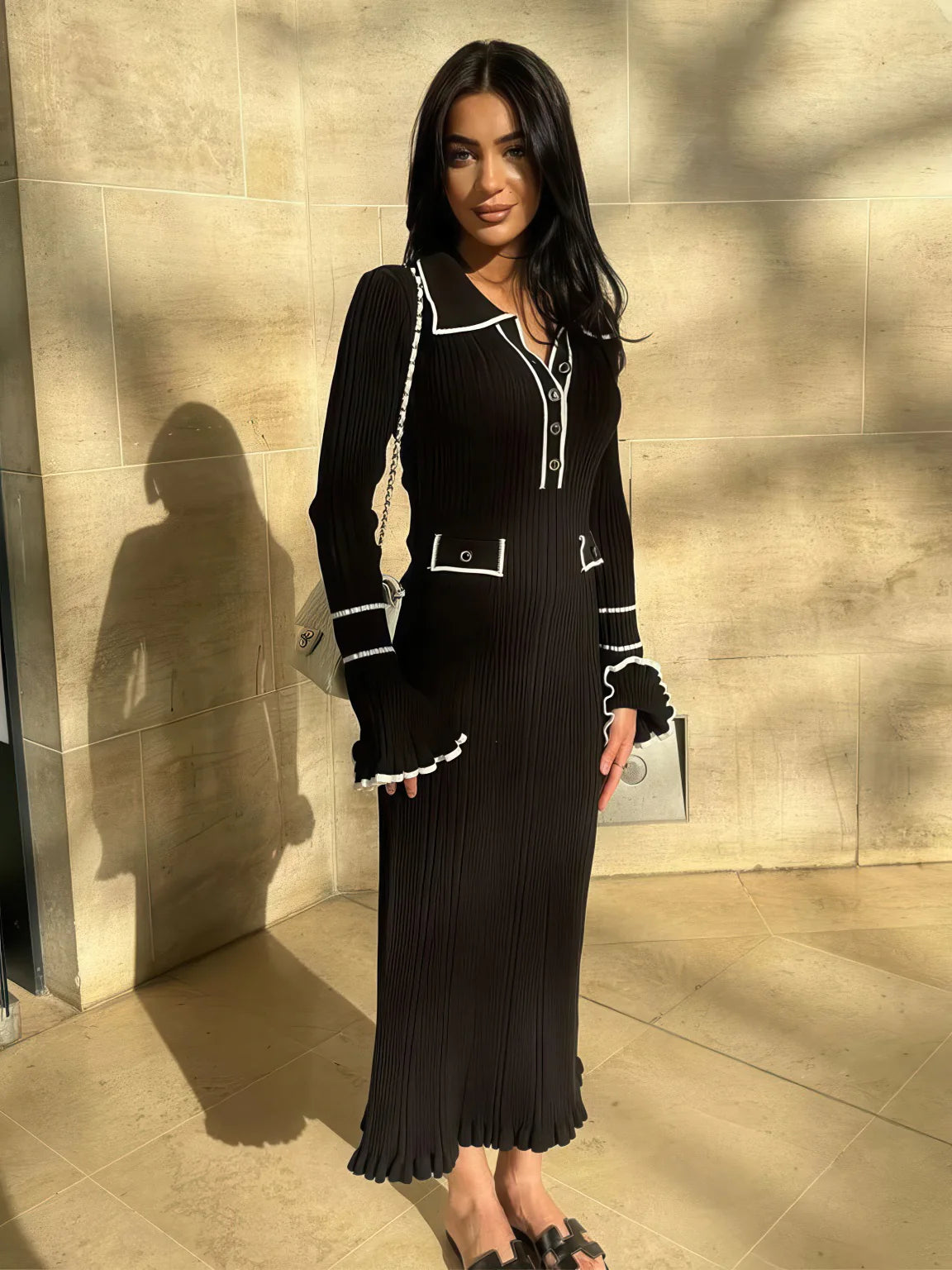 Woman in a black dress with white trim standing against a beige wall. #Color_Black