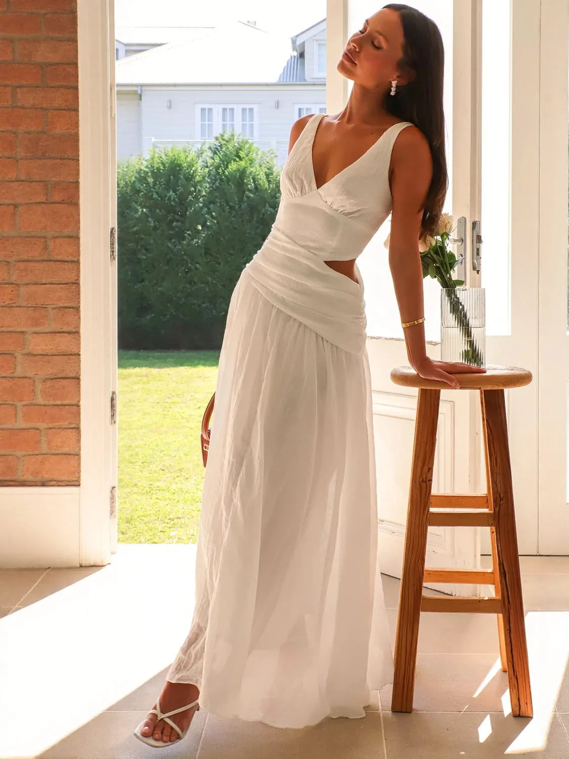 Woman in a white dress standing on a patio with a house and greenery in the background #Color_White