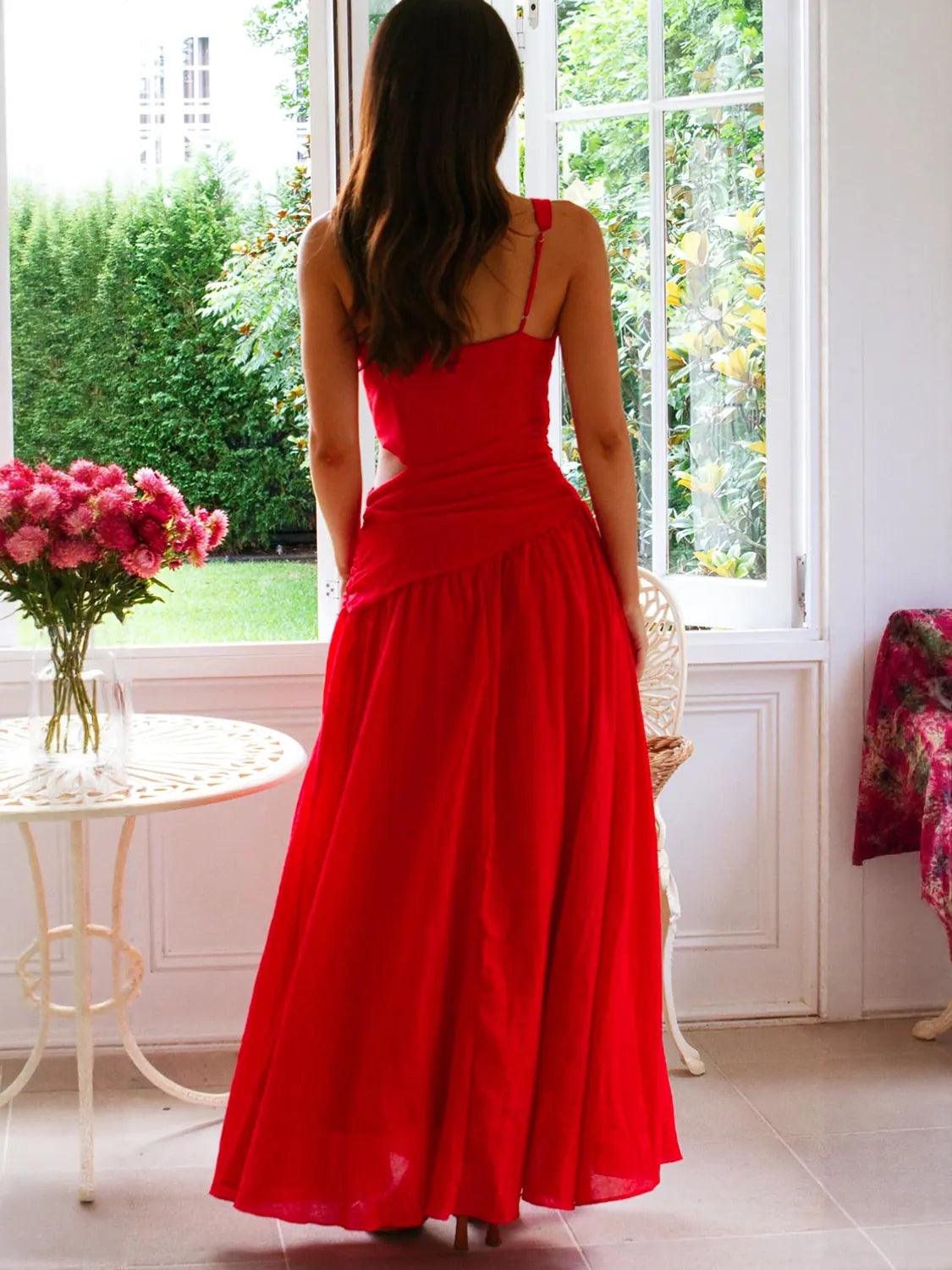 Woman in a red dress standing in a room with a window and flowers. #Color_Red
