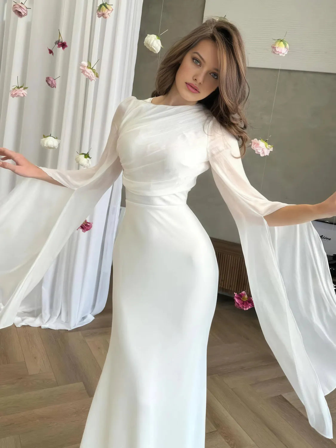 Woman wearing a white dress with sheer sleeves in a room with flowers. #Color_White