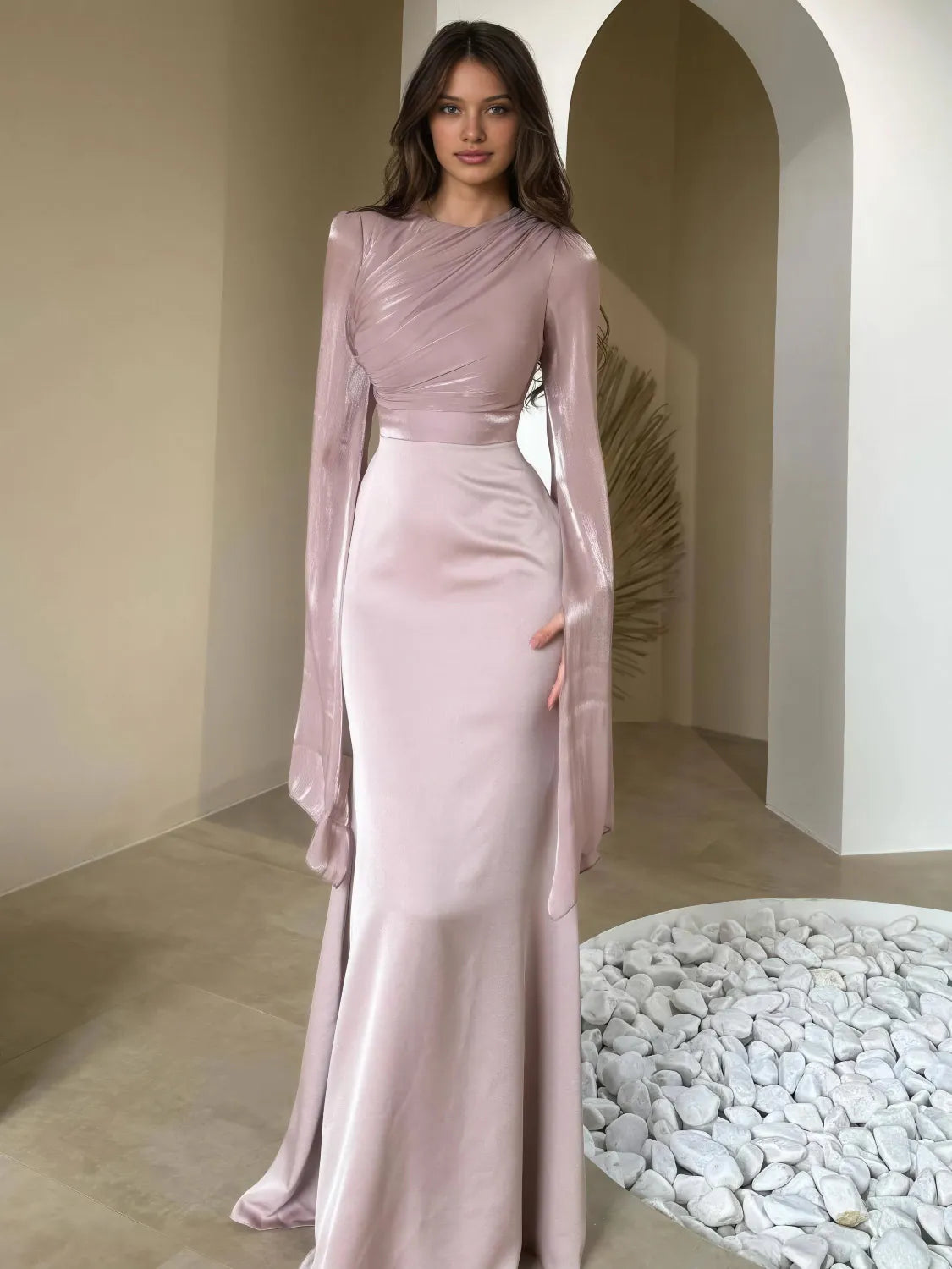 Woman wearing a long, light pink dress with a sheer overlay in an indoor setting. #Color_Pink