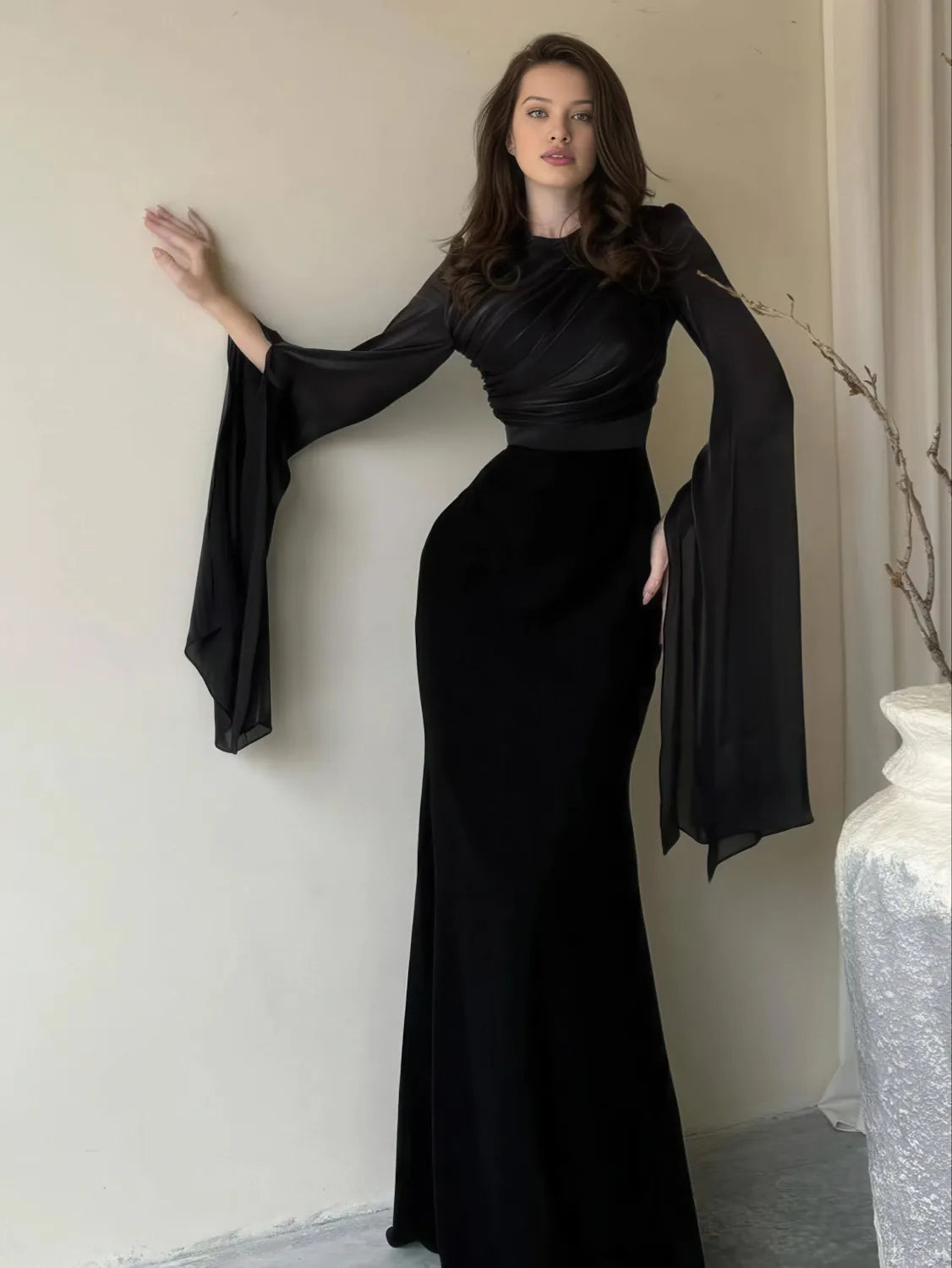 Woman wearing a black dress with flowing sleeves against a neutral background. #Color_Black