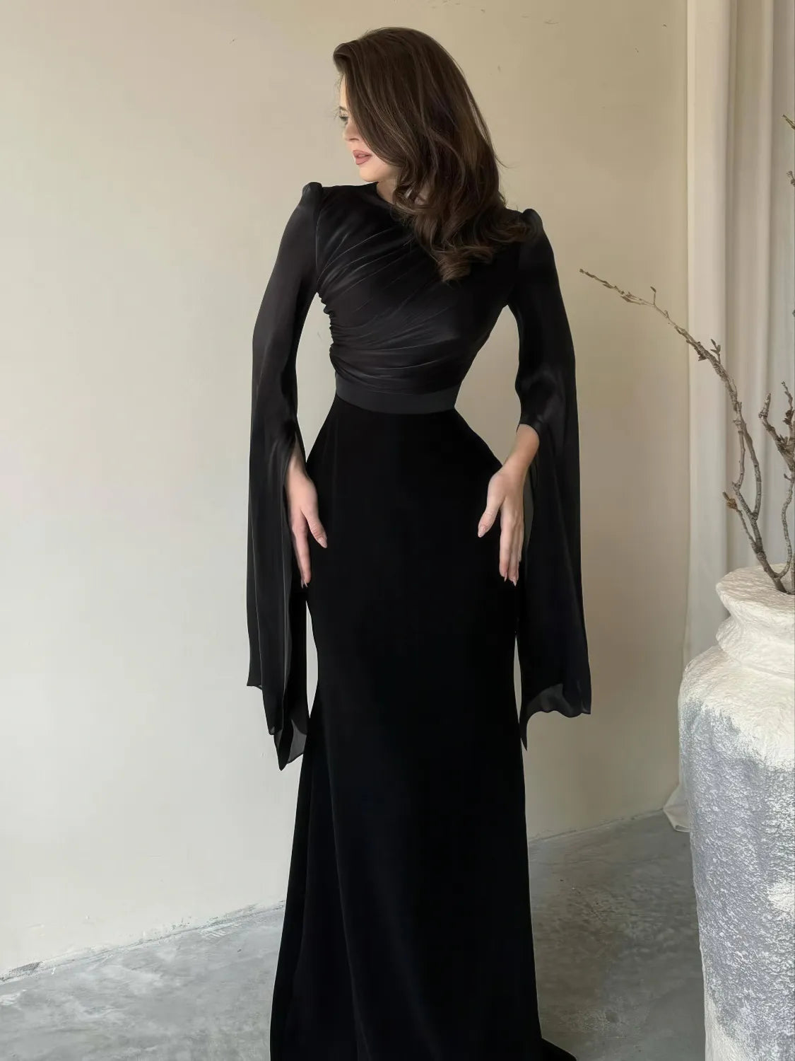 Woman wearing a black evening gown with cape-like sleeves in an indoor setting. #Color_Black
