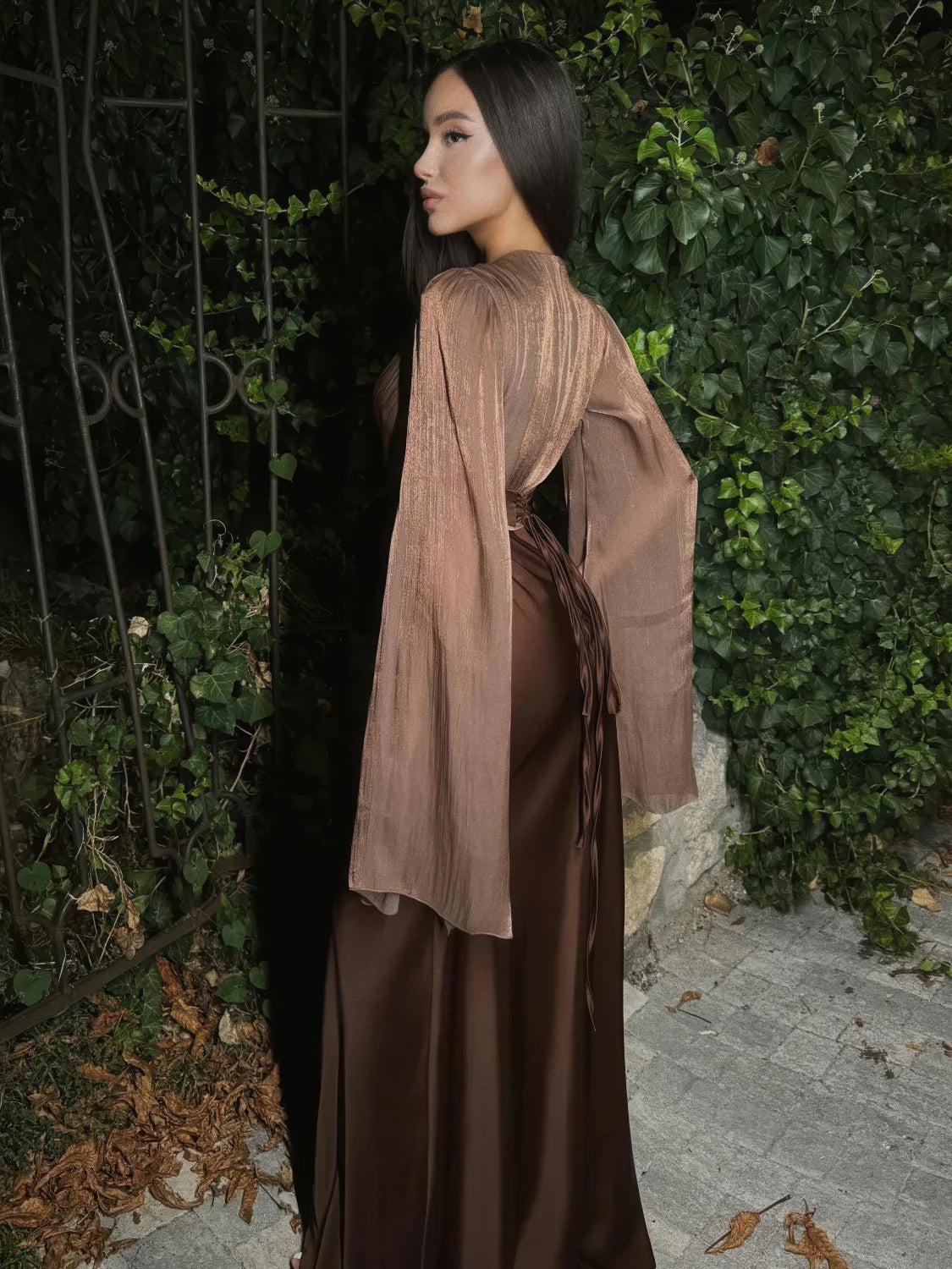 Woman wearing a brown dress with a sheer beige overlayer against a green foliage background. #Color_Coffee