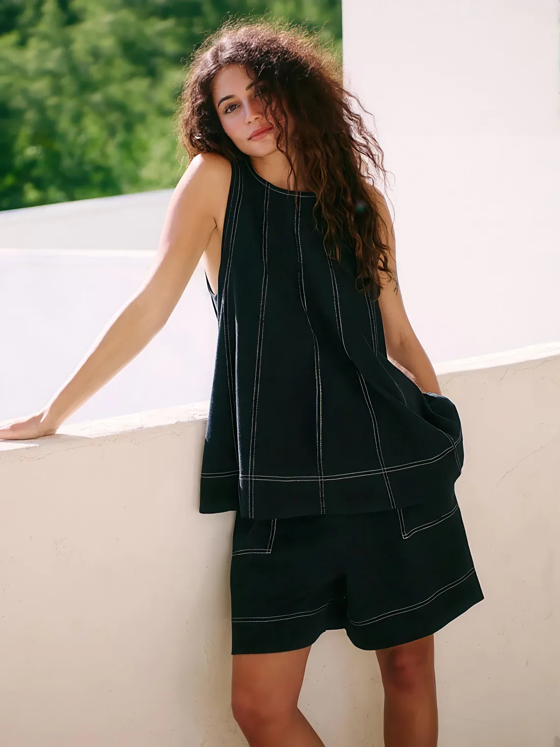 Woman wearing a black sleeveless top and shorts against a white wall with greenery. #Color_Black