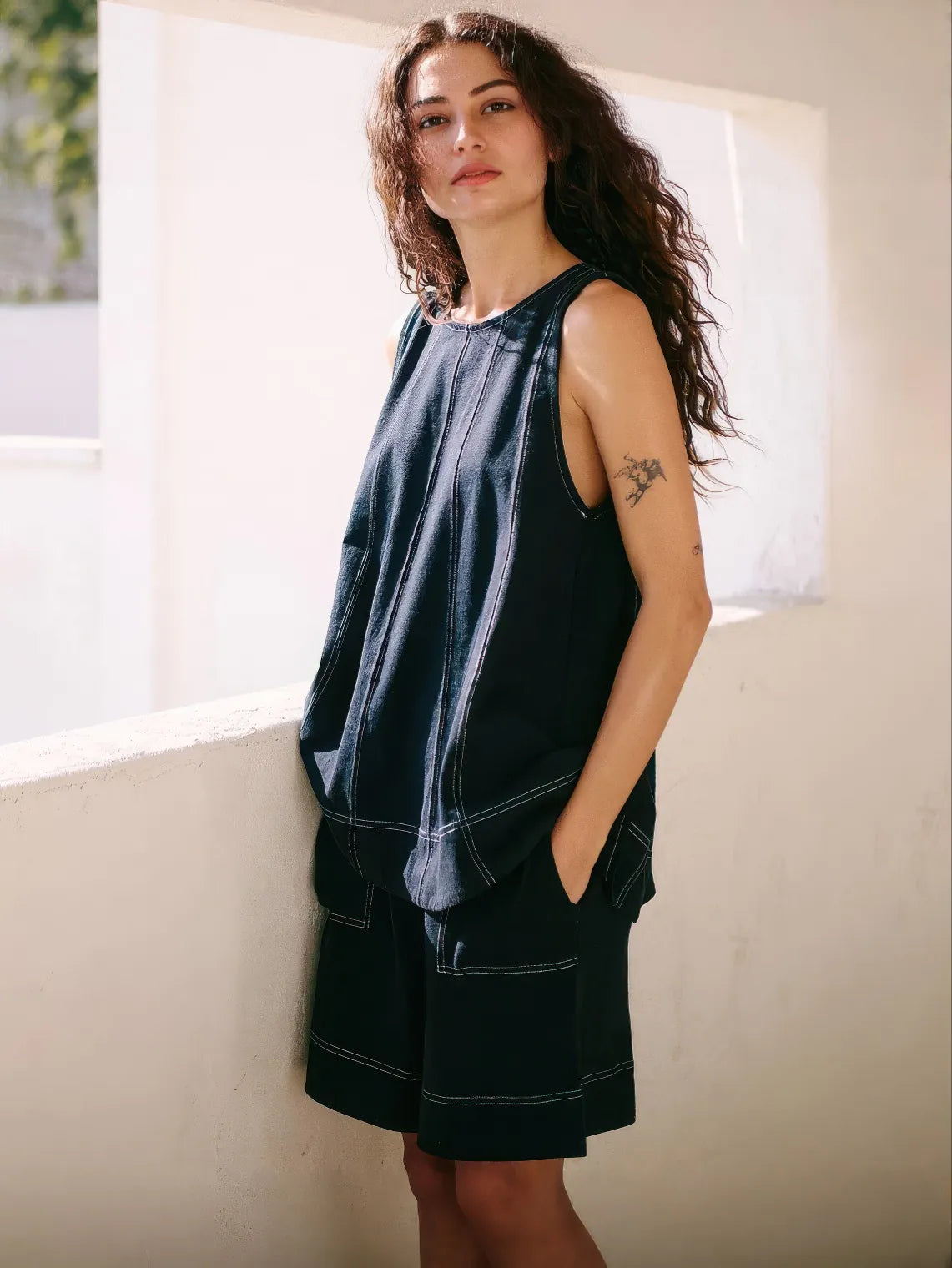 Woman wearing a sleeveless black top and black shorts standing against a white wall. #Color_Black