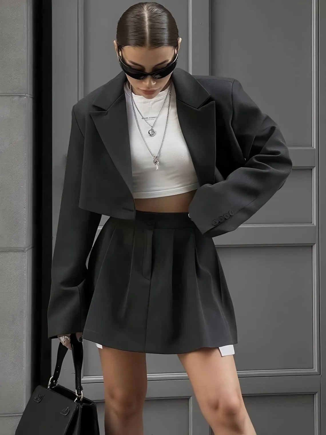 Person wearing a black blazer and shorts with a white top, standing against a gray wall. #Color_Black