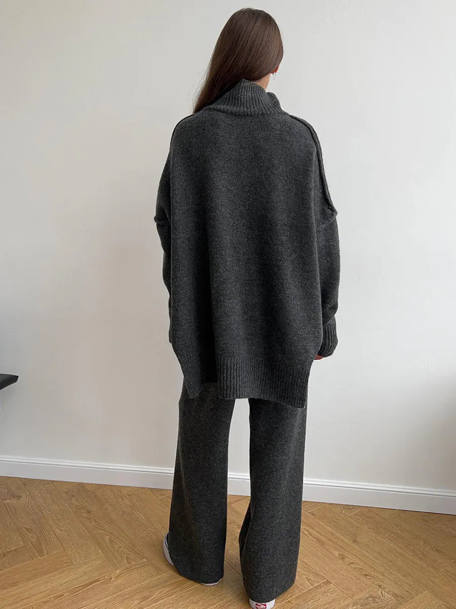 Person wearing a dark gray sweater and pants standing against a white wall. #Color_DimGray