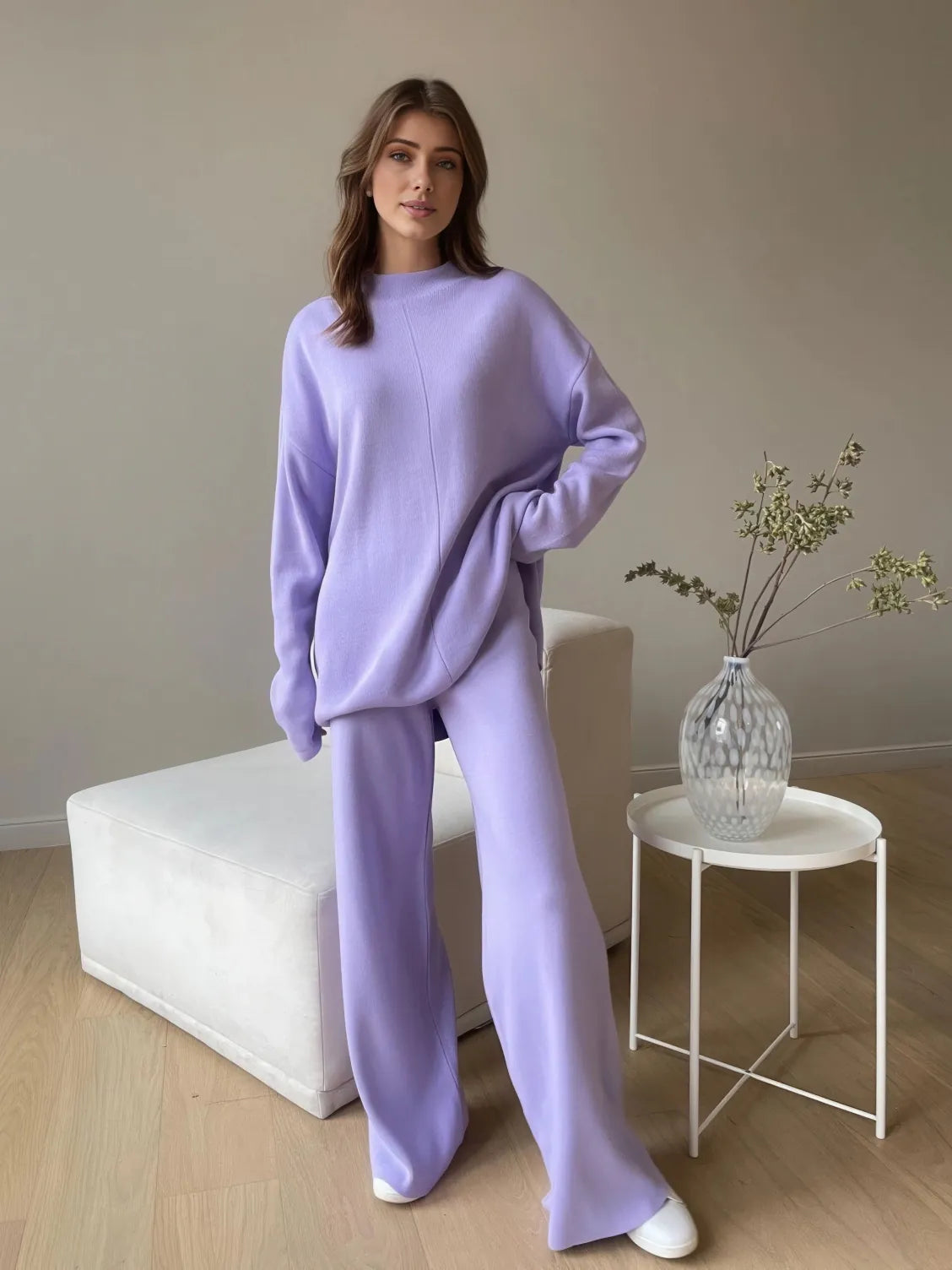 Woman wearing a lavender outfit standing in a minimalistic room with a vase on a side table. #Color_Purple