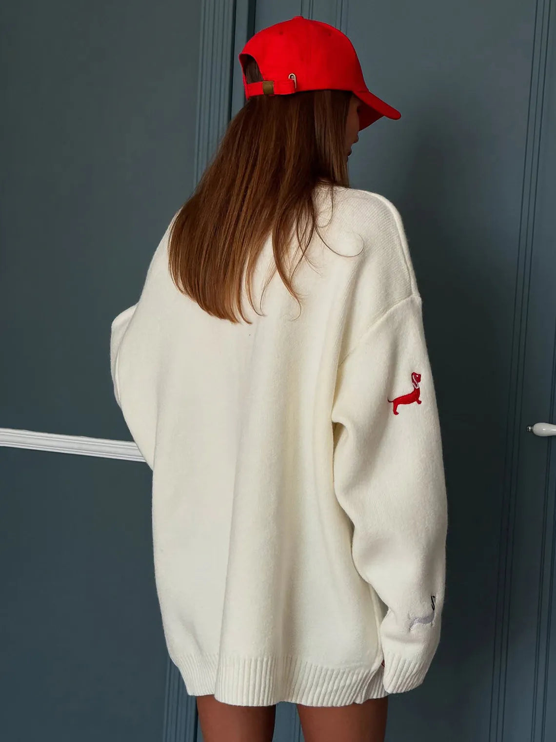 Person wearing a white sweater with a red dachshund design and a red cap against a gray wall. #Color_White