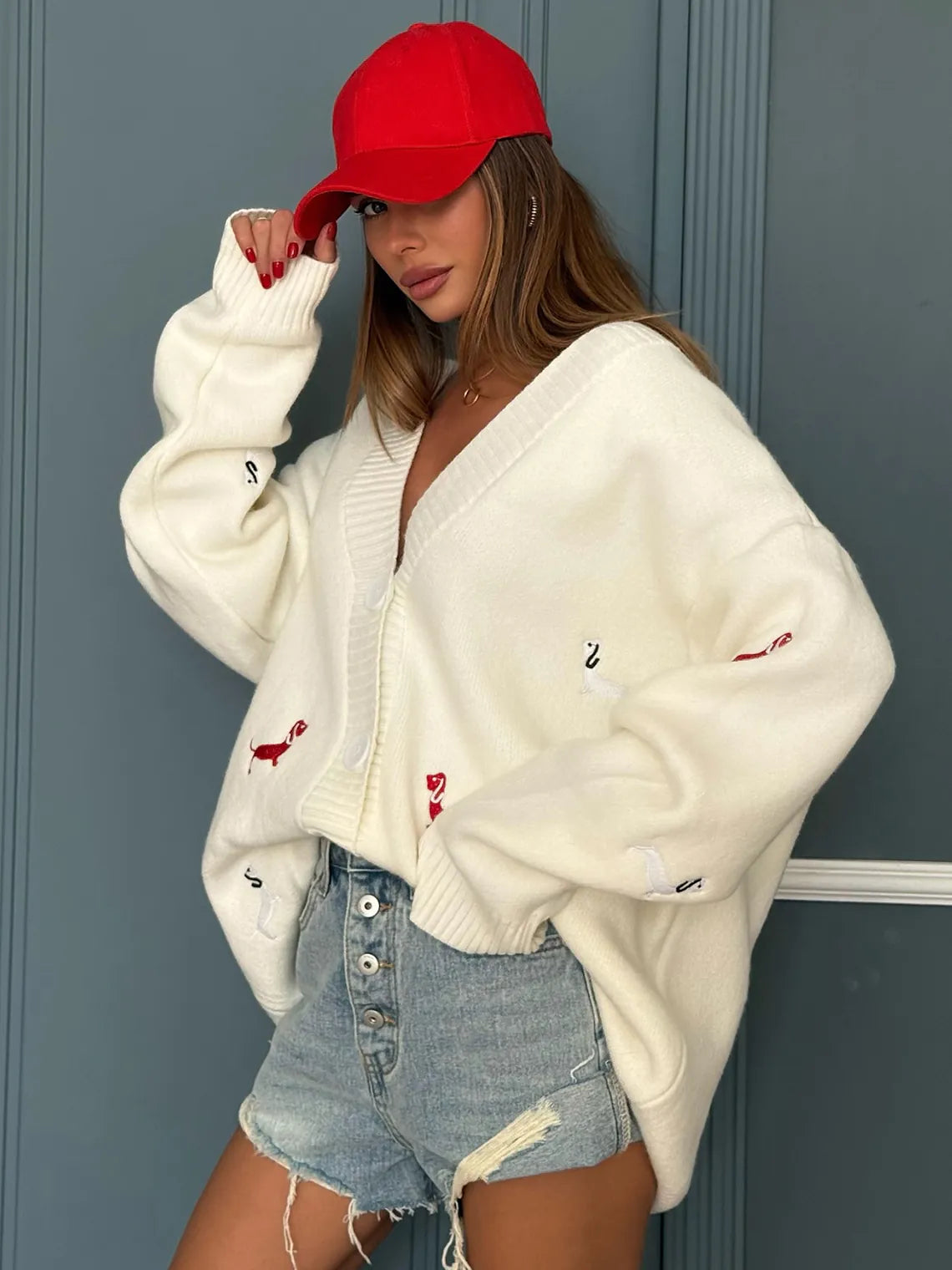 Person wearing a white cardigan with flamingo designs, denim shorts, and a red cap against a blue wall. #Color_White