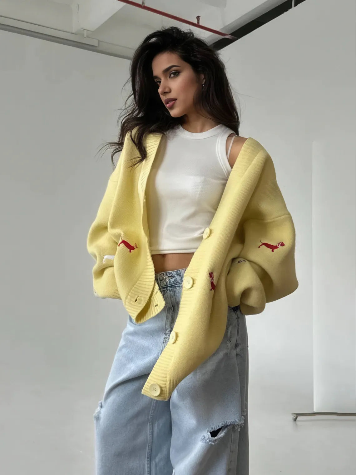 Woman wearing a yellow cardigan with red accents, white top, and blue jeans indoors. #Color_Yellow