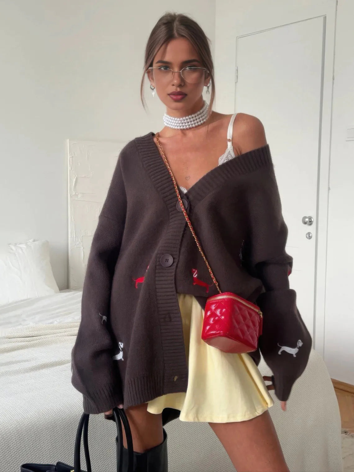 Woman wearing a dark brown cardigan with red patterns, a yellow skirt, and holding a red handbag in a bedroom setting. #Color_Brown