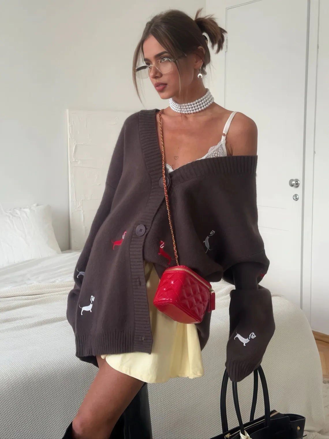Woman wearing a dark brown cardigan with dog embroidery, holding a red handbag, in a bedroom setting. #Color_Brown