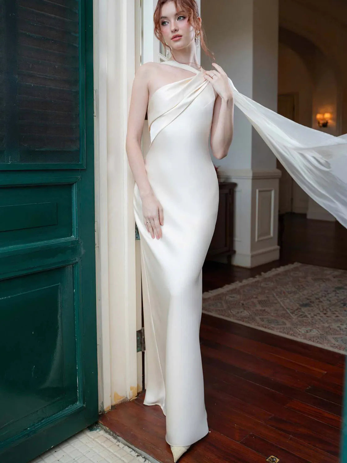 Woman in a white evening gown standing in a room with wooden floors and a green door. #Color_White