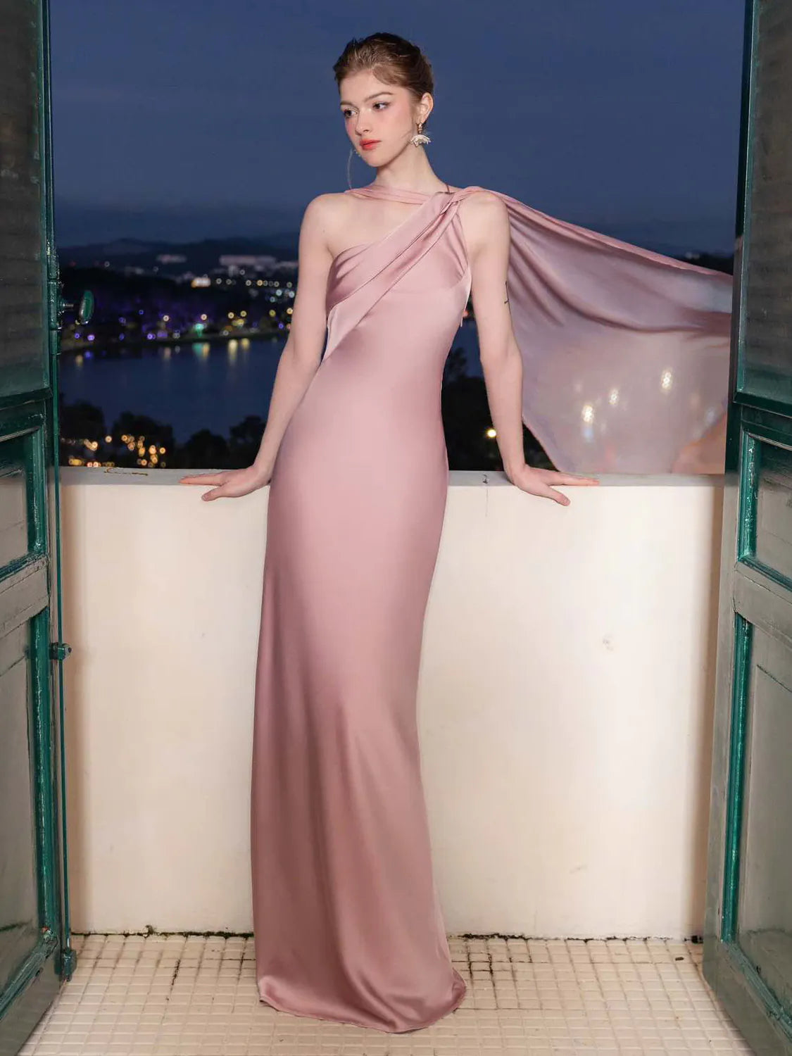Woman in a pink evening gown standing on a balcony with a night-time cityscape in the background. #Color_Pink