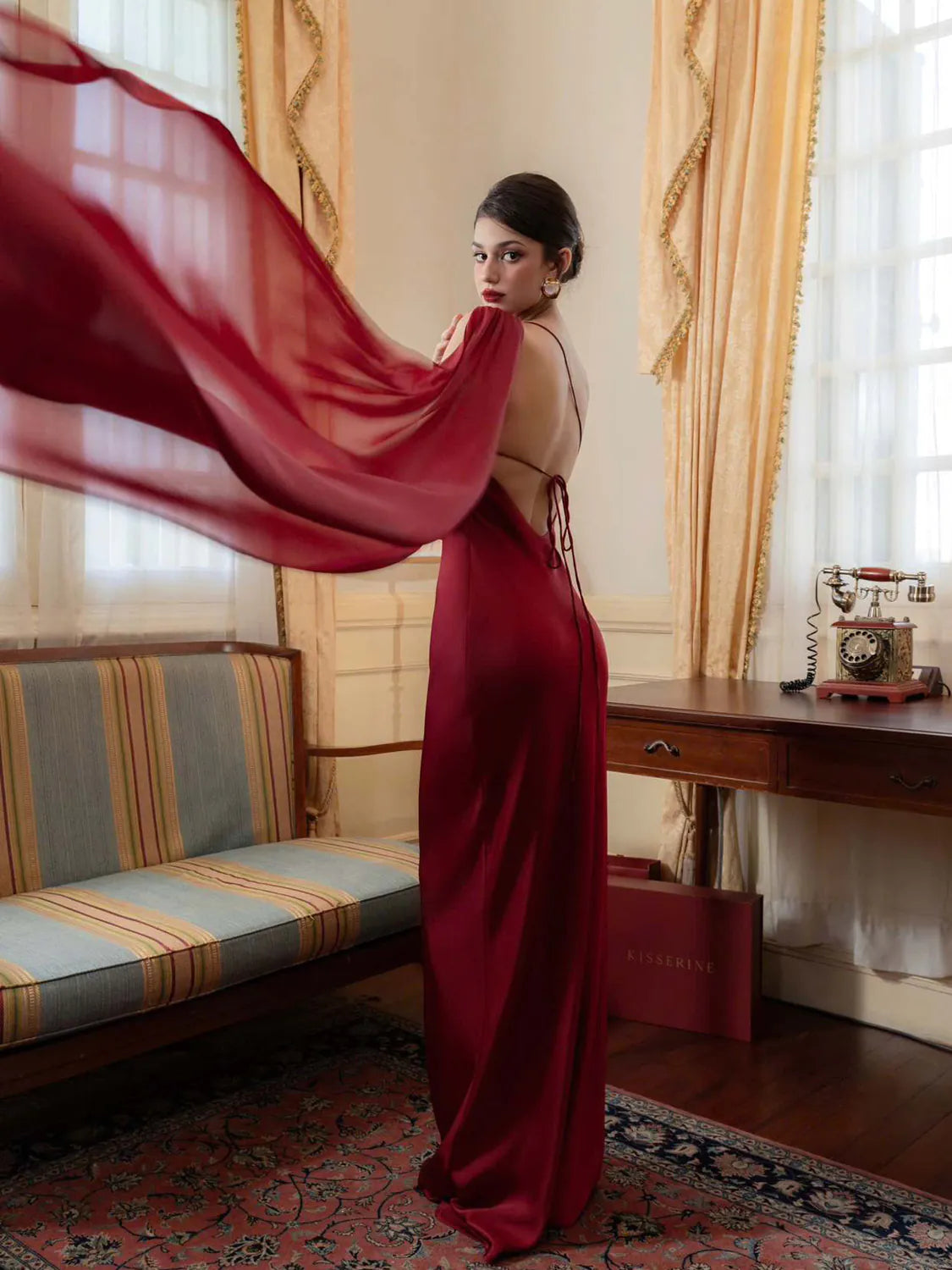 Woman in a red dress standing in a room with a desk and window. #Color_Red
