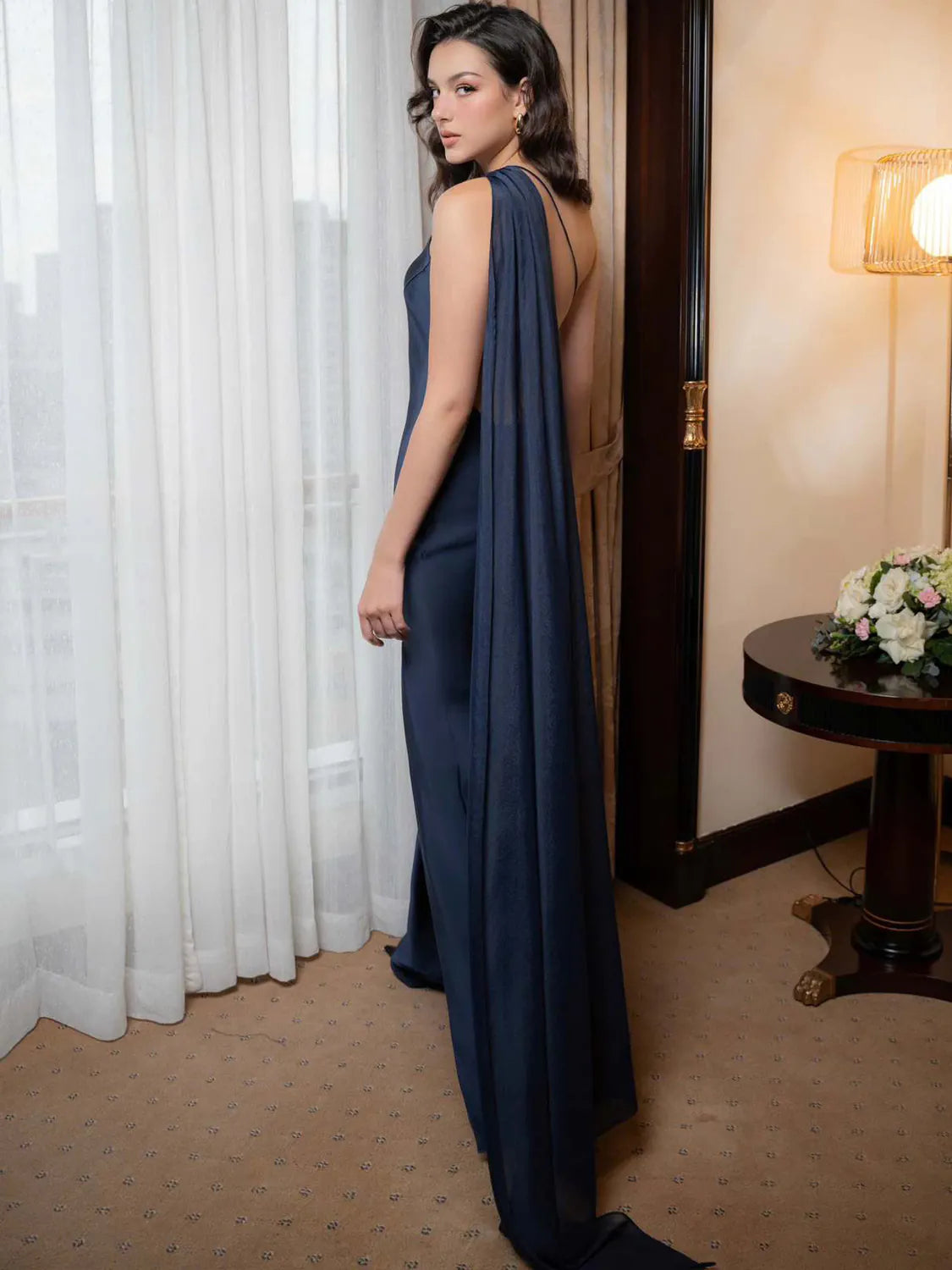 Woman in a navy blue evening gown standing in a room with white curtains and a table with flowers. #Color_DarkBlue