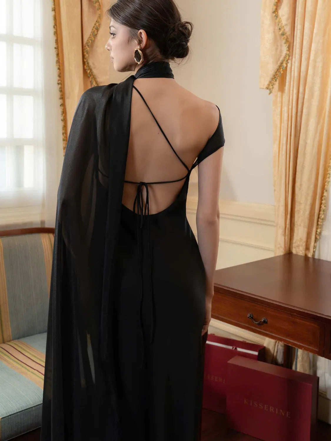 Woman in a black dress with a high slit standing in a room with ornate curtains and a red bag on the floor. #Color_Black