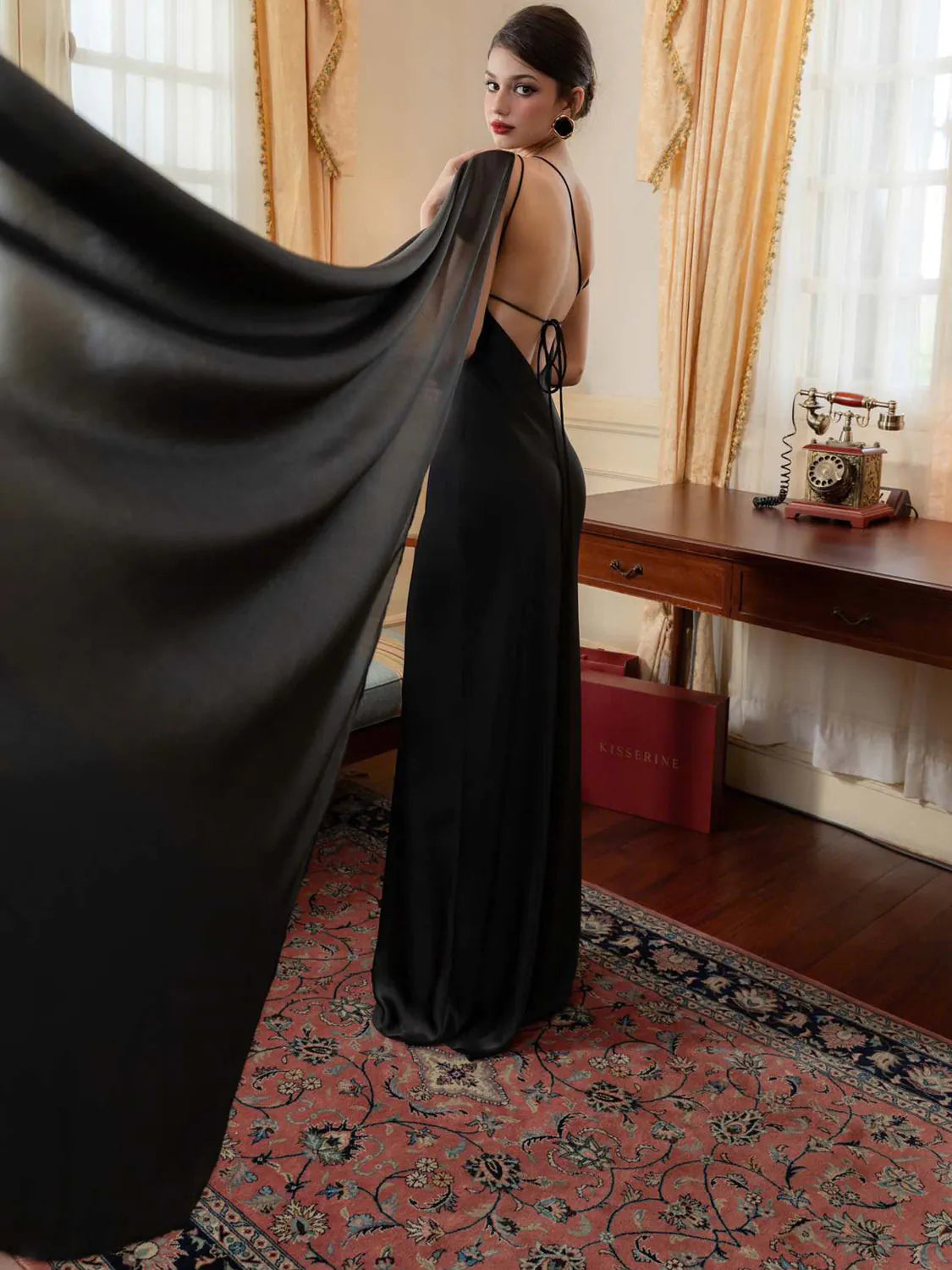 Woman in a black evening gown standing in an elegant room with a desk and decorative items. #Color_Black